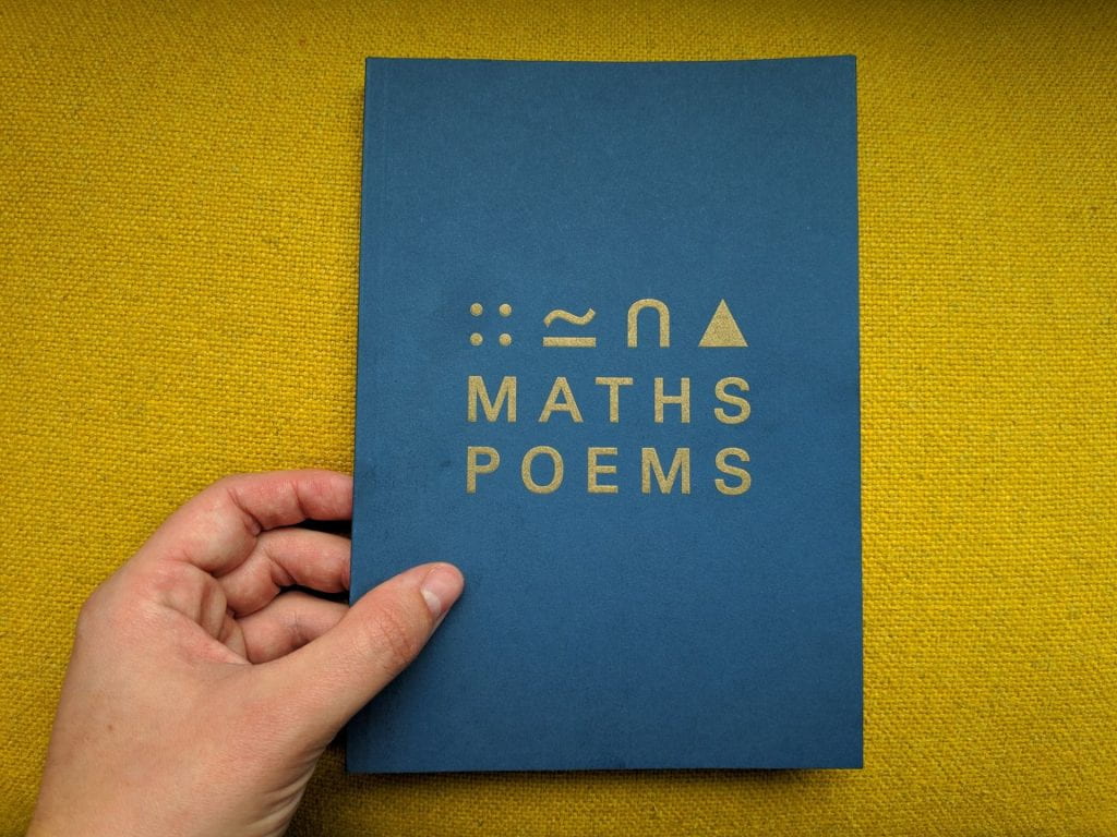 A image of a hand holding the "maths poems" anthology against a yellow backdrop. the book is blue with symbols and the words "Maths Poems" in Gold font.